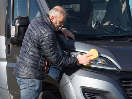 A male motorhome owner cleans his recreational vehicle front headlight with a sponge.Sunny dayの写真素材