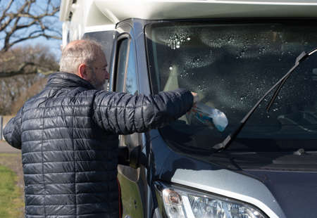 A bearded male motorhome owner cleans his recreational vehicle front windscreen window using a spray gun with blue liquid.Sunny dayの写真素材