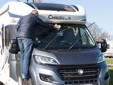 A male motorhome owner cleans his recreational vehicle front windscreen window .He stands on the tyre in order to reach.Sunny day. Chaussonのeditorial素材