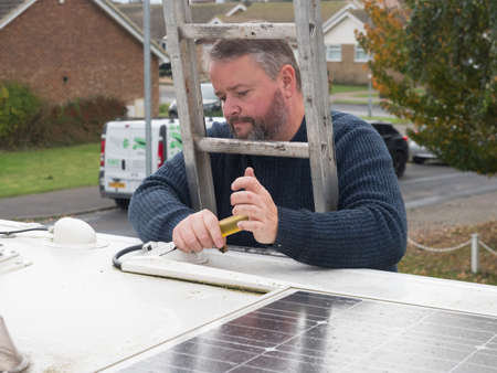 A man can be seen working from a ladder looking through rungs to remove plastic ducting from the roof top of a motorhome recreational vehicle.Solar panel visible.Viewed at roof top heightの写真素材