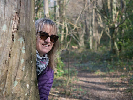 An attractive blonde lady wearing sunglasses, scarf and jacket peers round a tree trunk and smiles in a joking manner.A woodland rural countryside scene in backgroundの写真素材