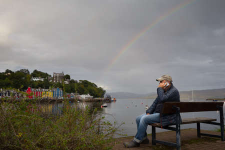 A man sits on a bench making a phone call on a mobile phone.He wears a baseball hat.In background is sea and coloured buildings of Tobermory in Scotlandの写真素材
