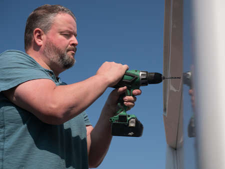 A male bearded mechanic uses a power drill to drill into a motorhome recreational vehicle.Shot from below with a blue sky backgroundの写真素材