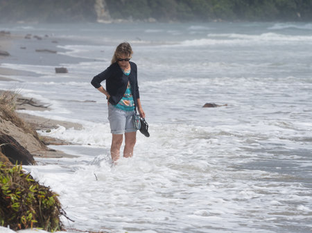 a female tourist stands in the surf water on a beach watching the waves cover her feet as she holds her sandals.Vacation,Recreationの写真素材