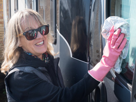 An attractive lady smiling at camera uses a cloth to wipe the side window of her motorhome recreational vehicle.She wears pink rubber gloves and sunglassesの写真素材