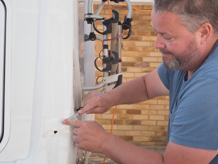 A bearded male mobile mechanic handyman concentrating and smiling as he uses a file to repair damage at the rear of a motorhome recreational vehicle.He wears a t-shirtの写真素材
