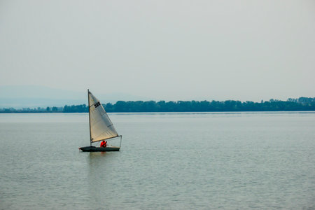 A woman is sitting in a sailing boat, steering and at the same time talking on the phone on a lake with a wooded shore in the backgroundの写真素材