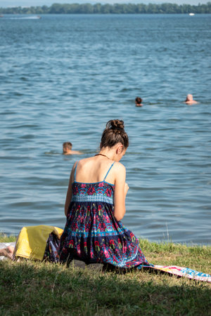 A beautiful young woman is sitting on a grassy beach by a lake in a summer dressの写真素材