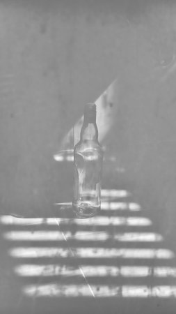 A moody black and white still life featuring a single empty glass bottle. Dramatic light streams in, casting strong, striped shadows across the surface, creating a captivating pattern of light and dark. This image evokes feelings of solitude, introspection, loneliness, or quiet contemplation.の写真素材