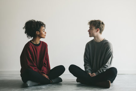 Two people are sitting on the floor, one wearing a red sweater and the other wearing a gray sweater. They are looking at each other and seem to be having a conversation. Scene is friendly and relaxedの素材