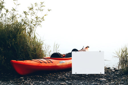 A red kayak is sitting on a rocky shore next to a white sign. The scene is peaceful and serene, with the water and the surrounding nature creating a sense of calmの素材