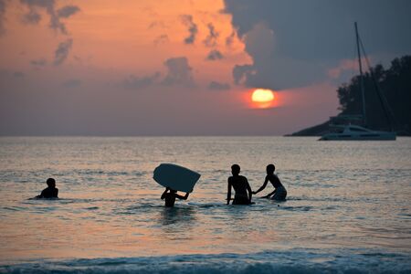 Silhouette of boys surfing on boards in ocean shore during sunsetの写真素材