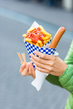 Female hands holding paper cone with a hot sausage and french fries with ketchupの写真素材