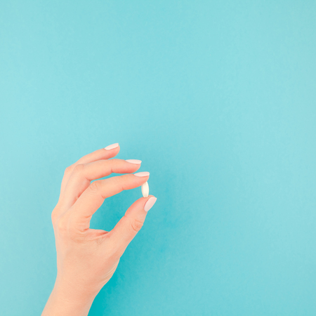 Woman hand holding a white pill on pastel blue paper background with copy space in minimal style, square template for text.の写真素材