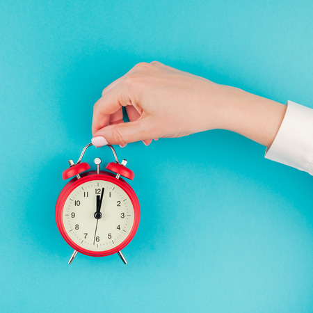 Creative flat lay concept top view of woman hand in white shirt holding the red vintage alarm clock on bright blue turquoise square color paper backgroundの写真素材