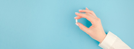Woman hand holding a white pill on pastel blue paper background with copy space in minimal style, template for text. Concept of medical treatment, pharmacy, healthcare. Long wide bannerの写真素材