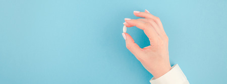 Woman hand holding a white pill on pastel blue paper background with copy space in minimal style, template for text. Concept of medical treatment, pharmacy, healthcare. Long wide bannerの写真素材