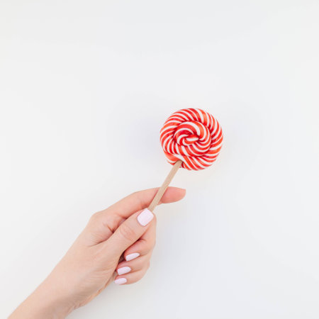 Woman hand with pastel manicure polish holding bright red swirling lollipop isolated on white background copy space minimalism style. Square Template for feminine social media. Fun celebration conceptの写真素材