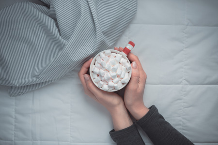 New Year or Christmas flat lay top view with Hot cacao coffee chocolate with marshmallows mug in woman hands Xmas holiday celebration in bed with lights. Concept blog social media 2019の写真素材