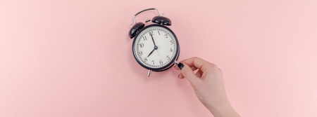Creative banner morning time emotions concept top view of woman hand holding black vintage alarm clock pastel millennial pink color paper background with copy space minimal style template for textの写真素材