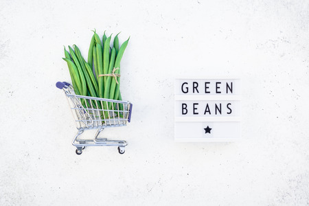 Creative scandinavian style flat lay top view of fresh green beans in shopping cart and text in lightbox on concrete table background copy space. Minimal house cooking concept promo templateの写真素材