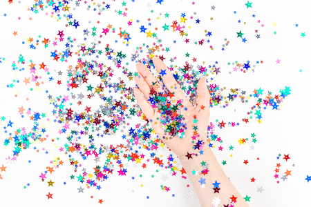 Bright festive color confetti star sparkles with woman hand on white background. Creative conceptual Christmas holiday birthday party top view flat lay backdrop for your blog, text or designの写真素材