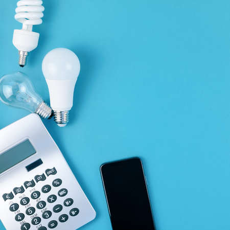 Energy and money saving and eco-friendly life in conceptual composition. Creative top view flat lay of new and old light bulbs, smartphone and calculator with copy space blue background minimal styleの写真素材
