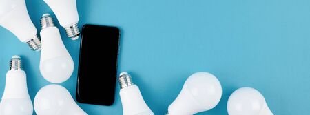 Energy-saving and eco-friendly life in conceptual frame pattern. Creative top view flat lay of LED light bulbs and modern smartphone mockup composition with copy space on blue background minimal styleの写真素材