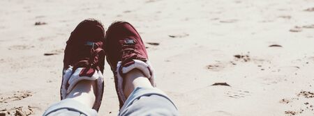 Feet of young woman in sport footwear and jeans relaxing after hiking on sand beach. POV. Adventure...