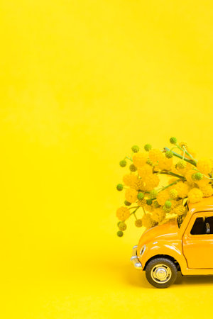 Moscow, Russia - February 23, 2019: 8 March International Women's Day card with toy model of retro car delivering bouquet of mimosa flowers on yellow background. Valentine's day, Flower delivery conceptのeditorial素材