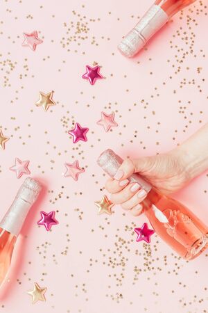 Christmas or New Year composition with bottle of rose champagne in woman hand and golden shiny sparkle star confetti on pastel pink background, top view. Celebration flat lay. Party creative conceptの写真素材