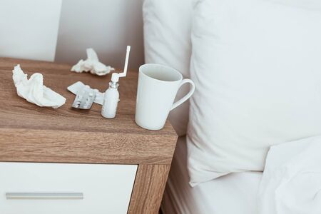Medicines, pills, nose drops, tissues and hot drink cup on bedside table in modern apartments. Being...