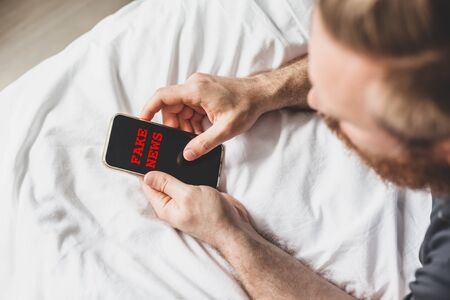 Fake news infodemics during Covid-19 pandemic concept. Man hands holding mobile phone reading global news or social net at home. Reading last news update in lockdown. Red text fake news on foregroundの写真素材