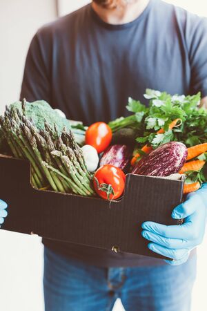 Fresh organic greens and vegetables delivery during coronavirus Covid-19 pandemic outbreak. Man hands wearing blue latex medical gloves holding box with safe grocery delivering in the house doorwayの写真素材