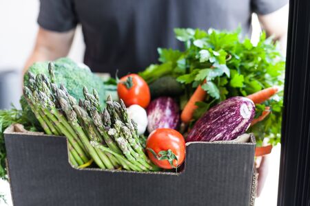 Fresh organic greens and vegetables delivery. Man hands holding box with farmer bio crop delivering in the house doorway. Small local business support. Online grocery shoppingの写真素材