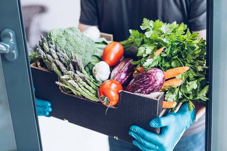 Fresh organic greens and vegetables delivery during coronavirus Covid-19 pandemic outbreak. Man hands wearing blue latex medical gloves holding box with safe grocery delivering in the house doorwayの写真素材