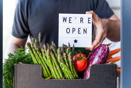 End of quarantine lockdown. Man holding lightbox with greeting text message We're open and Fresh greens and vegetables box. Welcoming grocery shop clients after coronavirus Covid-19 pandemic outbreakの写真素材