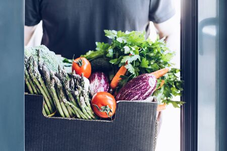 Fresh organic greens and vegetables delivery. Man hands holding box with farmer bio crop delivering in the house doorway. Small local business support. Online grocery shoppingの写真素材