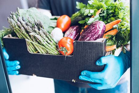Fresh organic greens and vegetables delivery during coronavirus Covid-19 pandemic outbreak. Man hands wearing blue latex medical gloves holding box with safe grocery delivering in the house doorwayの写真素材