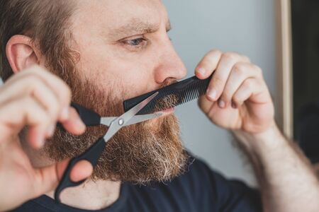DIY Selfcare during quarantine isolation. Adult handsome caucasian man cutting moustache and beard himself personally with scissors at home. Coronavirus outbreak and people shutdown, stay home conceptの写真素材