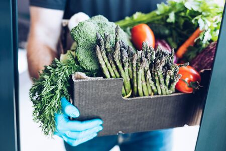 Fresh organic greens and vegetables delivery during coronavirus Covid-19 pandemic outbreak. Man hands wearing blue latex medical gloves holding box with safe grocery delivering in the house doorwayの写真素材