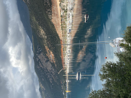 Marina with sailboats and reflections on calm water, surrounded by mountains under cloudy sky....