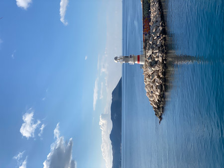 Lighthouse on rocky coastal jetty with calm sea and blue sky, ideal for serene backgrounds. Mobile photoの写真素材