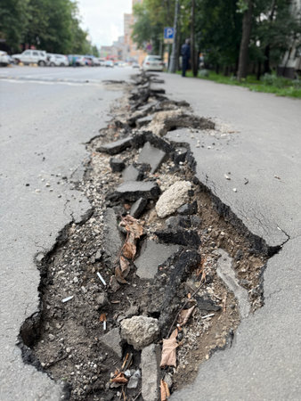 Damaged urban road with large crack showing broken asphalt and exposed dirt with leaves. Mobile...