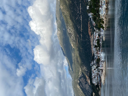 Mountain landscape with coastal town, sea, and dramatic clouds in scenic panoramic view. Mobile...