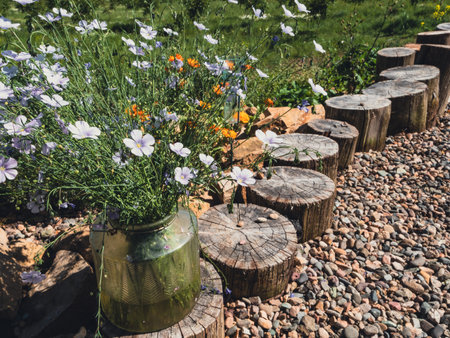A glass vase filled with white flowers rests on a wooden log in a natural setting. The log path leads through a bed of gravel with other blooming flowers in the background.の写真素材