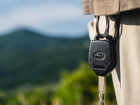 A black car key, featuring the Subaru logo, hangs from the waistband of a persons pants, with a blurred background of lush green hills. Kojori, Georgia - June 14, 2024.のeditorial素材
