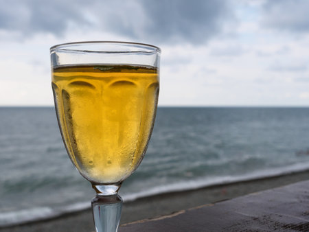 A glass filled with chilled white wine stands against a backdrop of the ocean. The tranquil waves and overcast sky create a calming, relaxing atmosphere.の写真素材