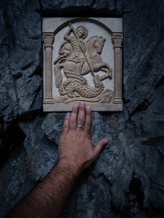Serene moment of mans hand reaching out to connect with the Saint George icon intricately set into the natural rock wall, reflecting a deep spiritual reverence and historical religious significance.の写真素材