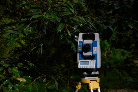 Terengganu, Malaysia - January 20, 2025 : A professional total station surveying instrument, mounted on its tripod, stands prominently in a lush, green outdoor environment. The bright sunlight illuminates the details of the precision equipment, including its control panel and various sensors. The natural, leafy background suggests a setting for land surveying, construction site preparation, or mapping projects in the field.の写真素材